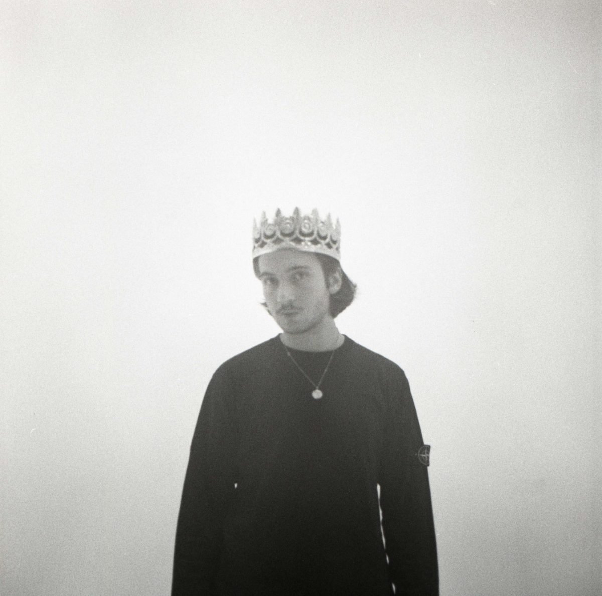 Jeune homme portant une couronne et un collier, en noir et blanc, regarde vers la gauche, avec un fond simple et clair.