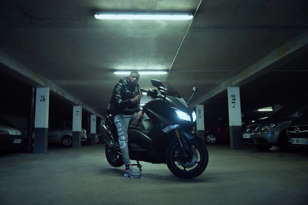 Un homme debout à côté d'une moto dans un parking souterrain, voitures en arrière-plan.