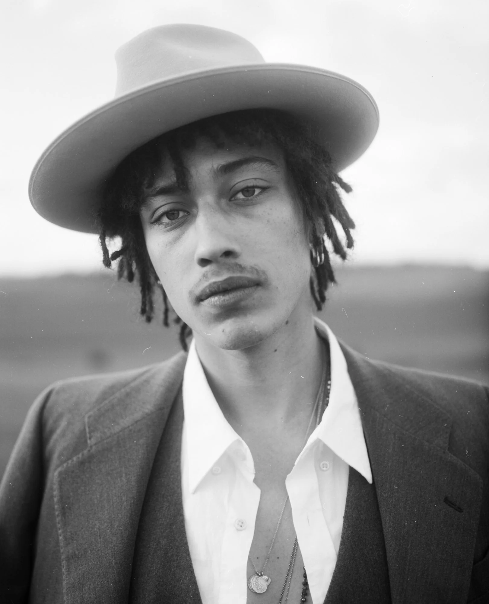 Portrait en noir et blanc d'une personne portant un chapeau ample et un blazer, avec une chemise blanche, portant plusieurs colliers, un regard sérieux et décontracté.