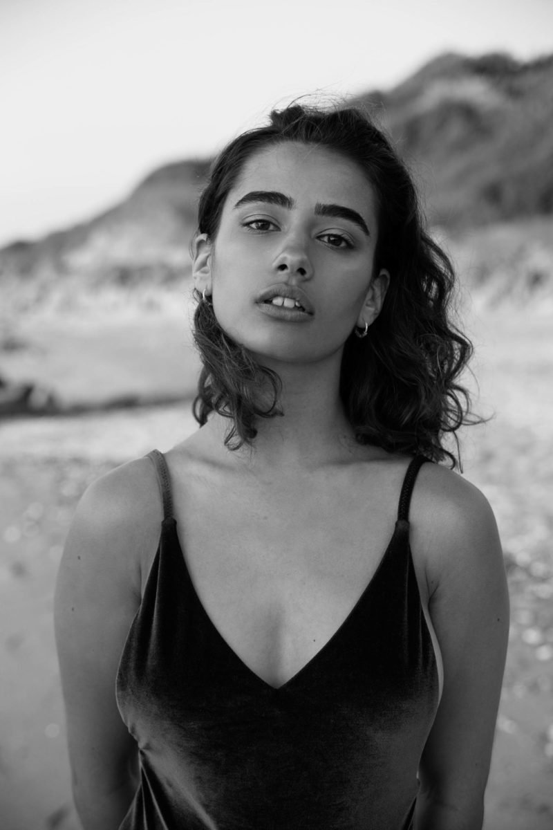 Une jeune femme en débardeur noir posant sur la plage avec un décor rocheux en arrière-plan.
