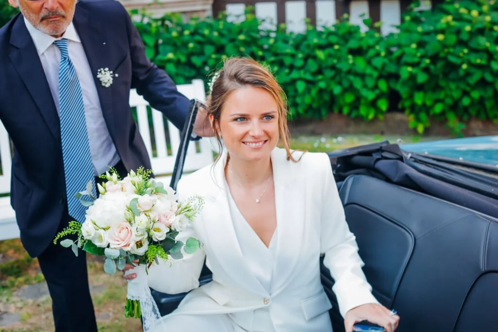 Une femme souriante en robe blanche assise dans une voiture en train d'être déposée, tenant un bouquet de fleurs, avec un homme en costume à côté.