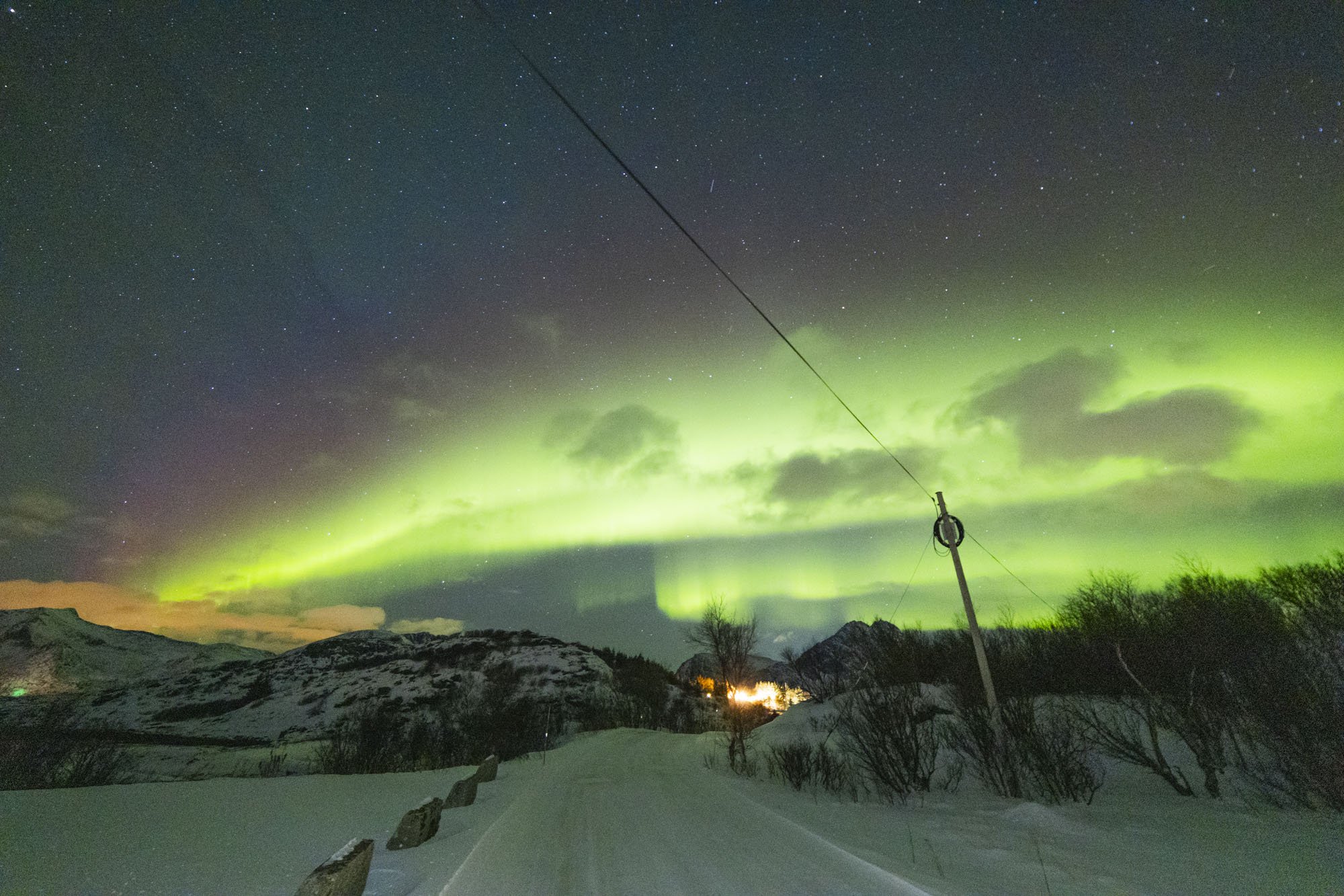Ciel étoilé avec aurore boréale verte en dessous, vue d'une route enneigée, montagnes au loin, et un poteau électrique à tromso