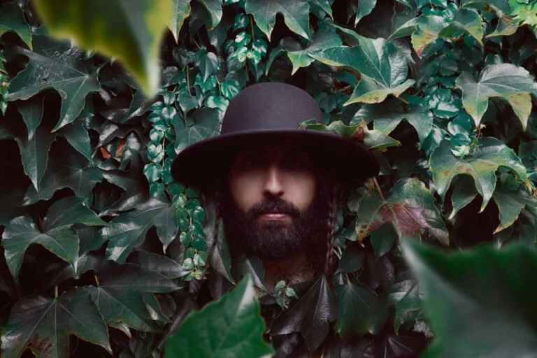 Une personne portant un chapeau noir et un manteau noir, entourée de feuilles de lierre vertes.