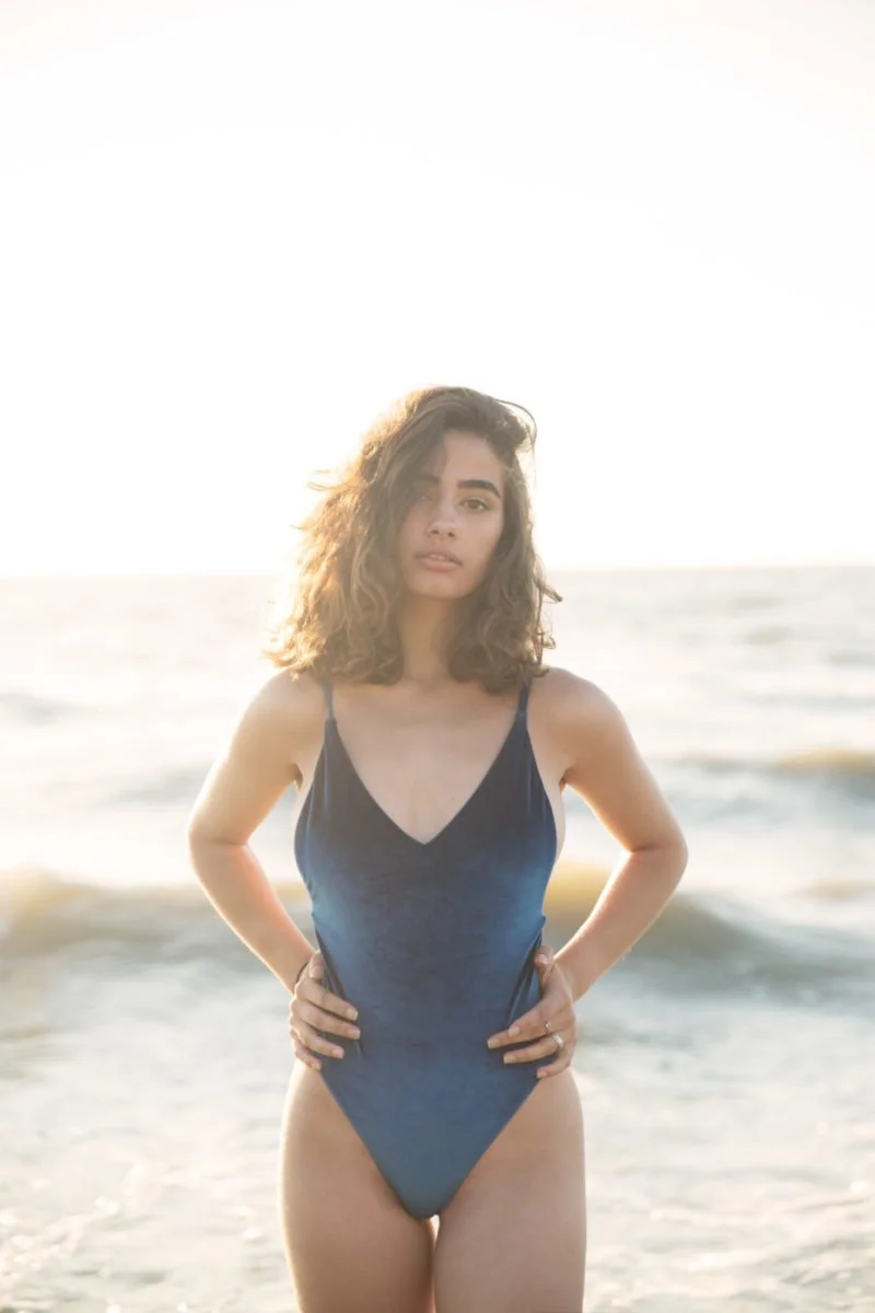 Jeune femme en maillot de bain bleu, posant sur la plage avec la mer en arrière-plan, lors d'un coucher de soleil.