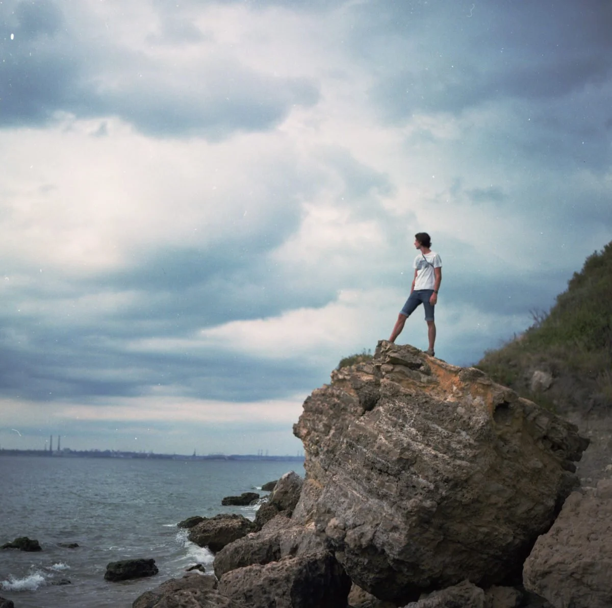 Jeune garçon debout sur une grosse roche au bord de la mer, regardant vers l'horizon, avec un ciel nuageux en arrière-plan.