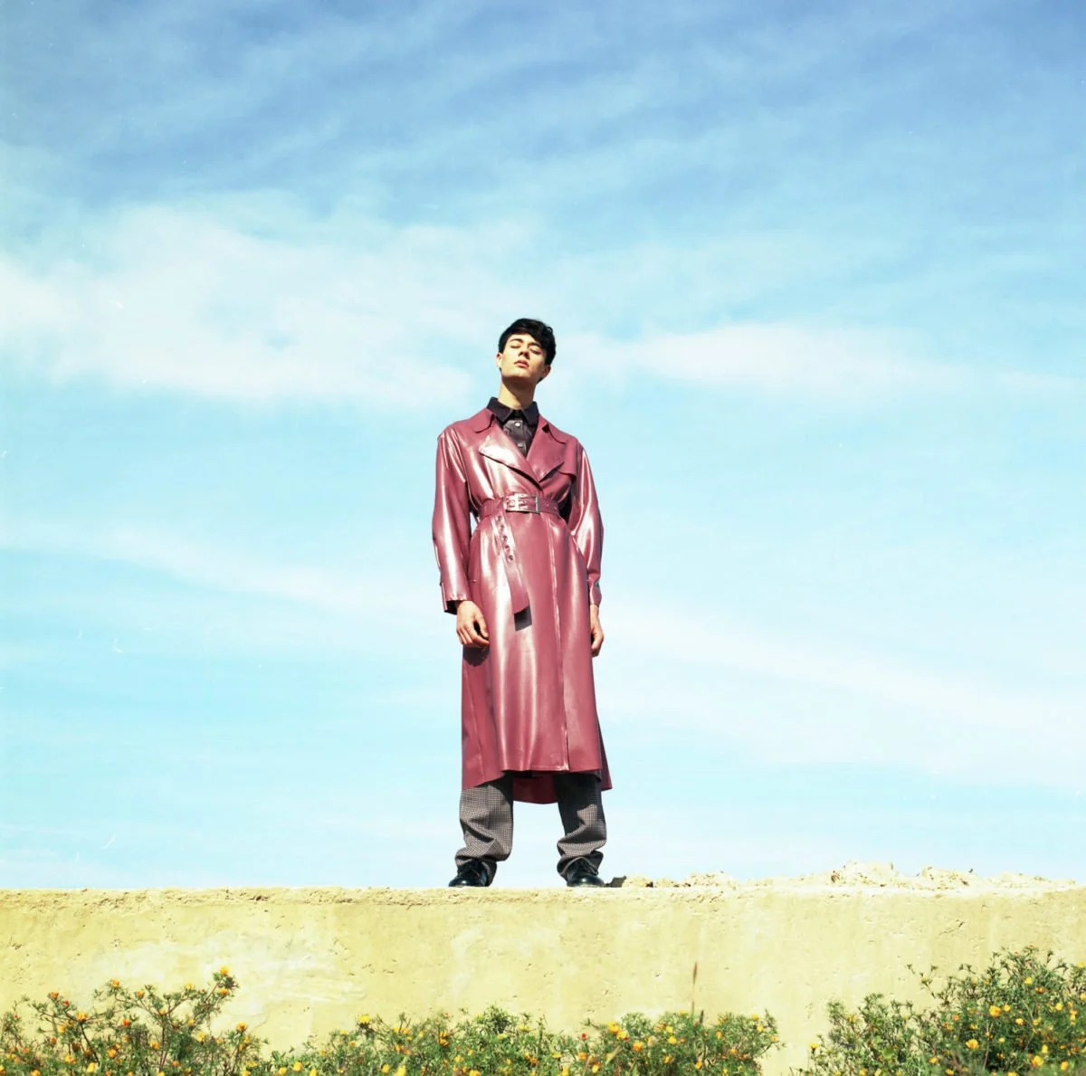 Une personne debout sur une plateforme en terre, portant un manteau long en vinyle rose et un pantalon à carreaux, sous un ciel bleu clair avec quelques nuages. Arthur Avellano