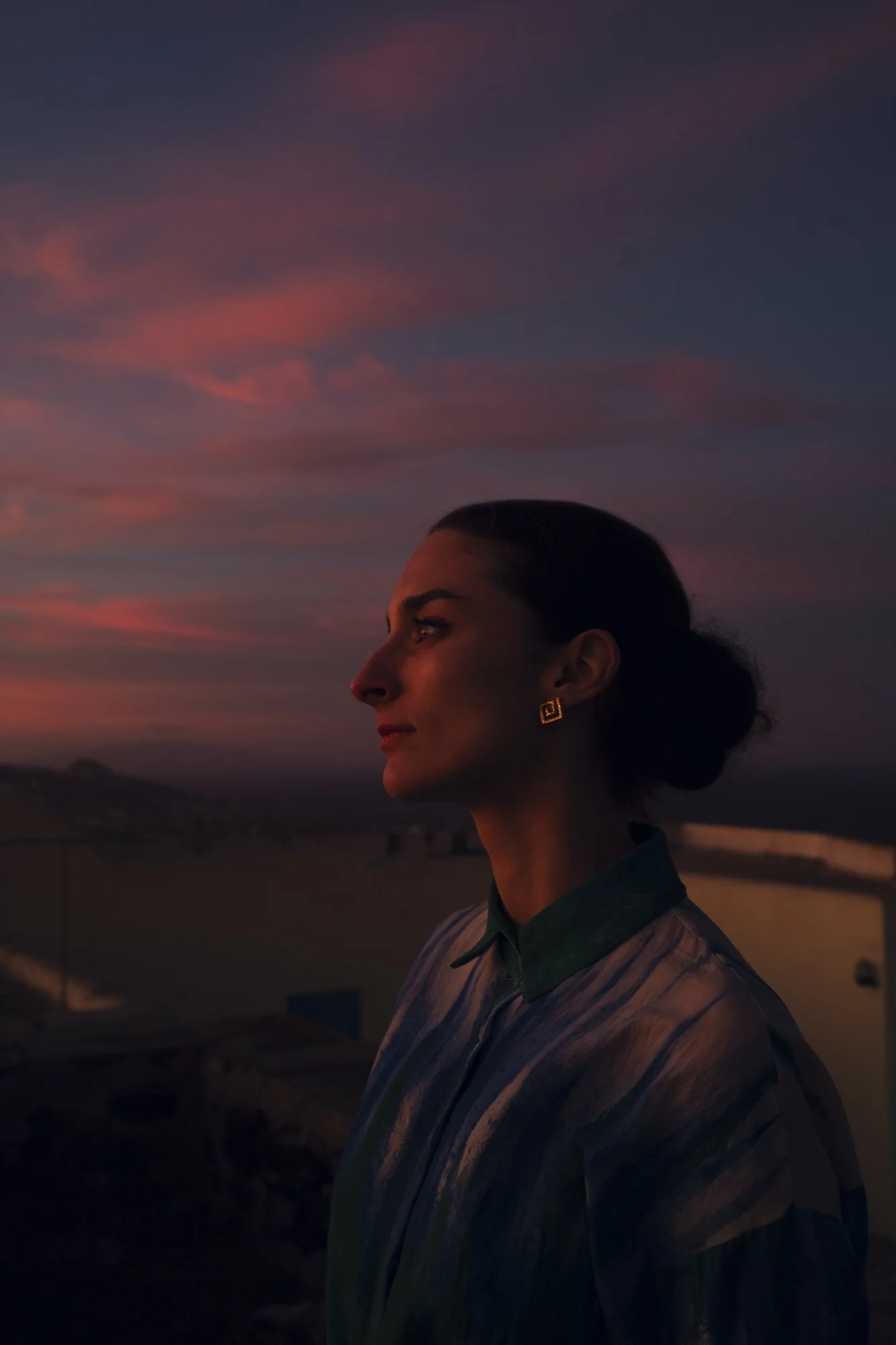 Profil d'une femme avec boucle d'oreille géométrique, regardant vers la gauche, sous un ciel crépusculaire rose et violette à santorin