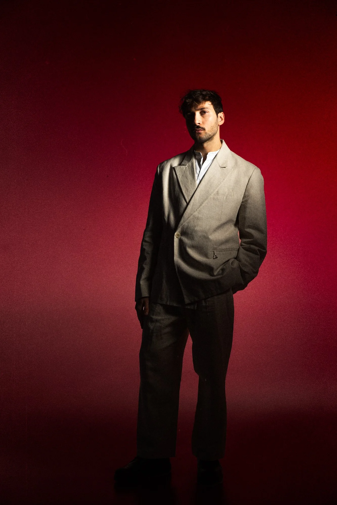 Un homme porte un costume beige avec une chemise blanche, debout contre un fond rouge foncé, en éclairage dramatique.