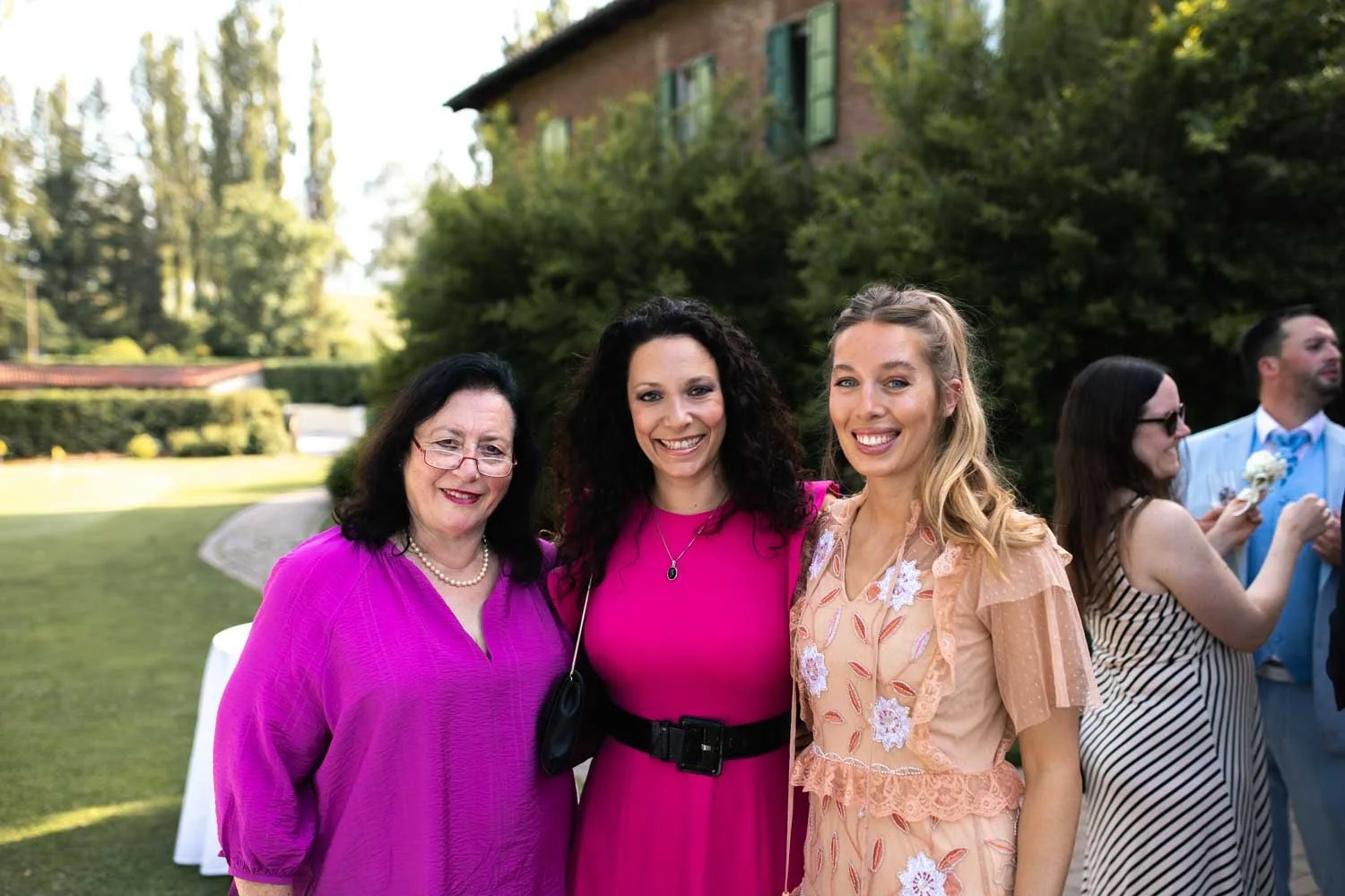 Trois femmes souriantes posant ensemble lors d'un événement en plein air, avec d'autres personnes en arrière-plan, dans un jardin ou un parc. Seules trois sont clairement visibles au premier plan.
