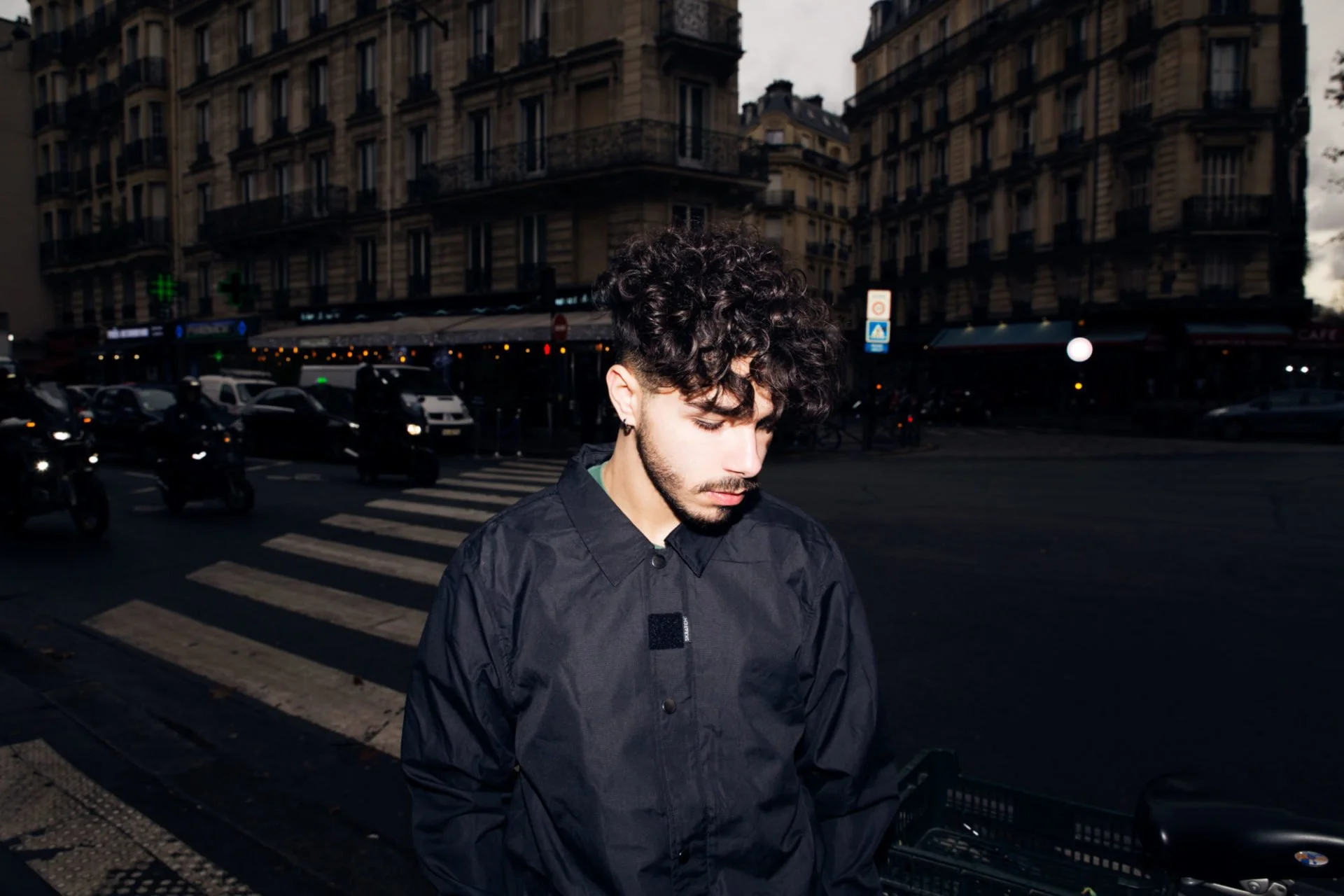 Jeune homme aux cheveux bouclés et barbe, porte un blouson noir, regarde son téléphone dans une rue urbaine en soirée ou au crépuscule.