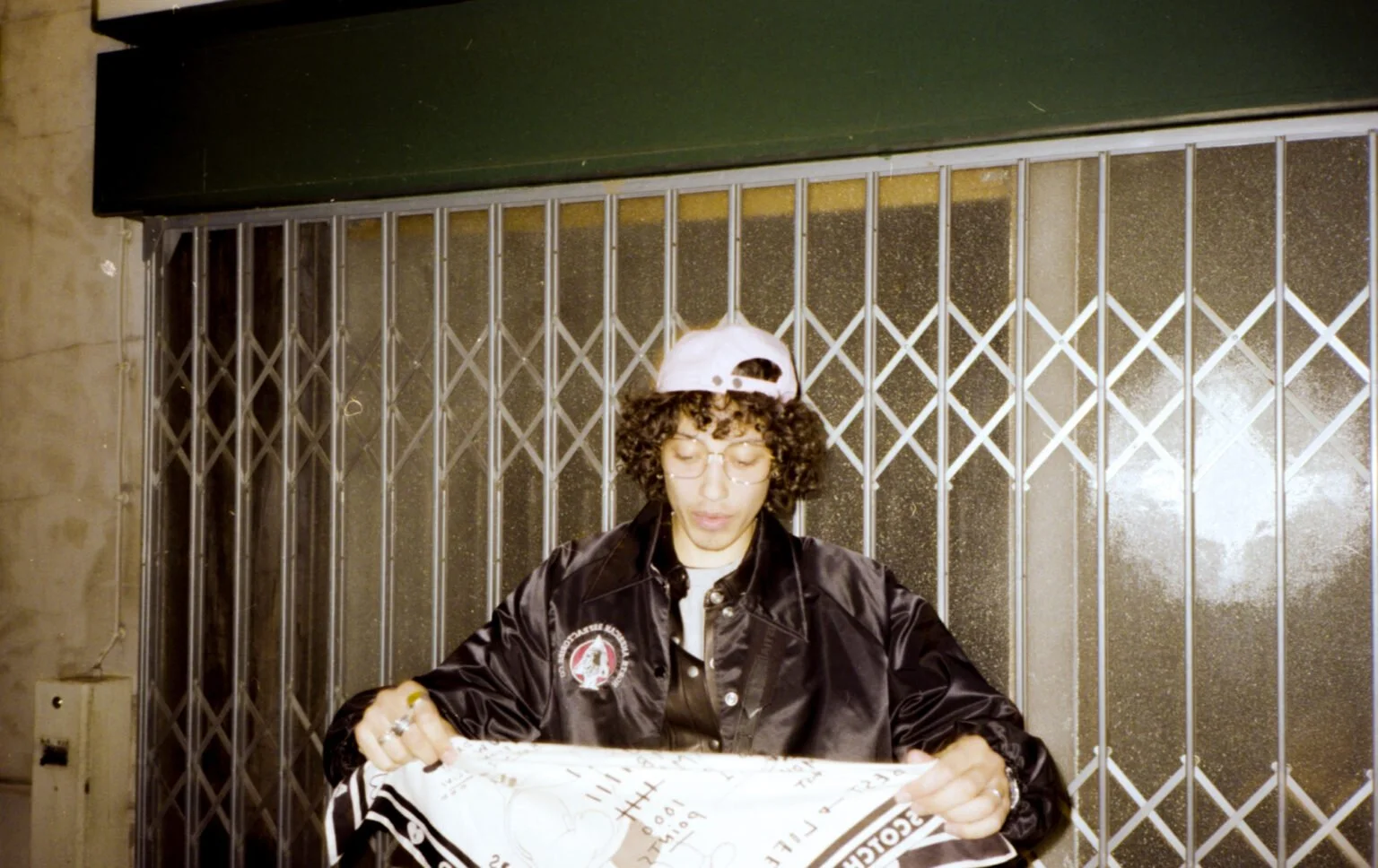 Une personne portant une casquette blanche, des lunettes, une veste en satin noir, et tenant un journal, assise devant une grille métallique.