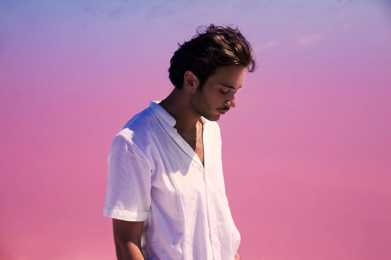Un jeune homme avec des cheveux bruns, portant une chemise blanche, regarde vers le sol avec un fond de ciel de coucher de soleil rose et violet.