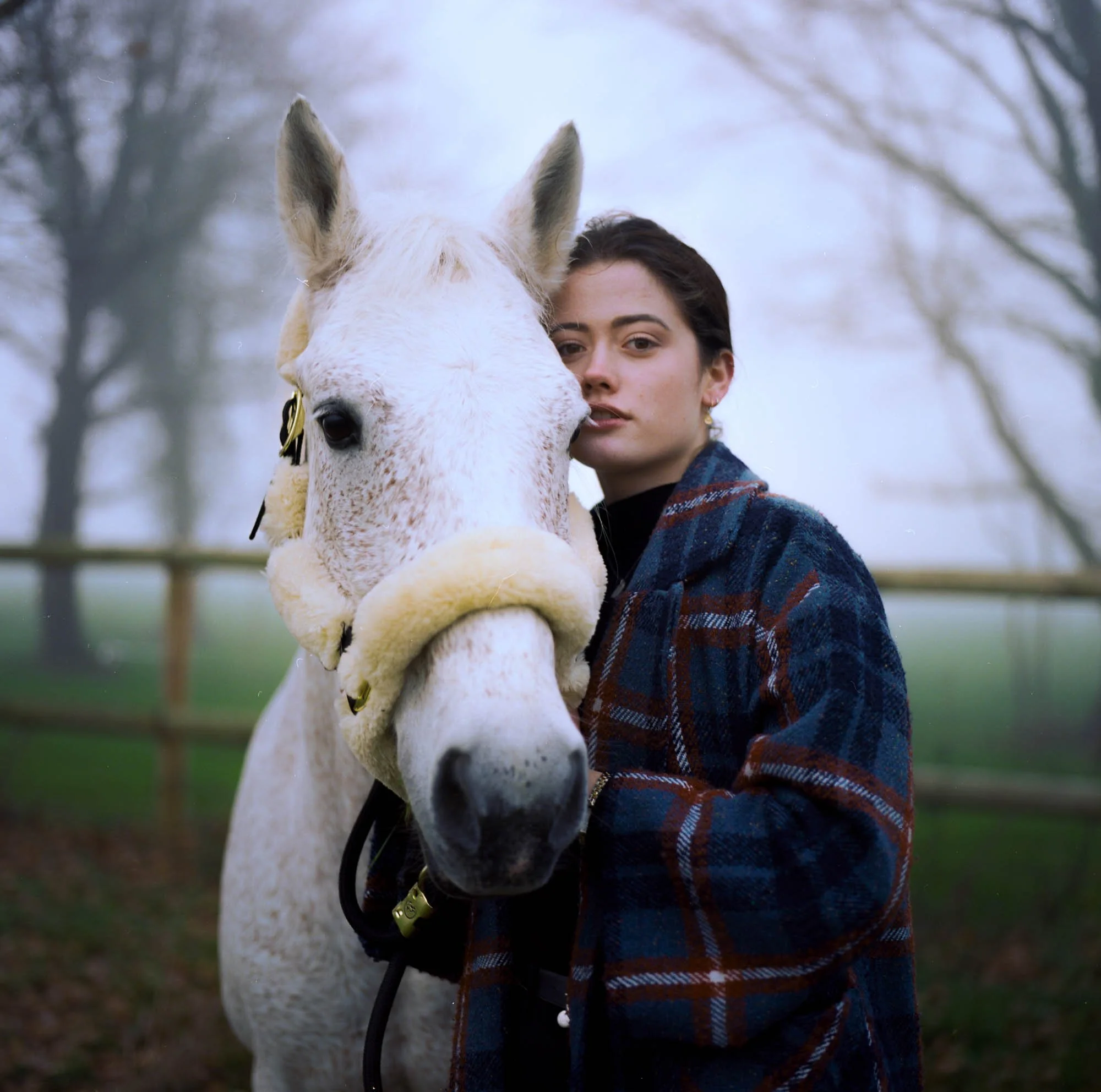 Une femme avec un pull à carreaux, tenant en laisse un cheval blanc dans un espace extérieur avec des arbres en arrière-plan.