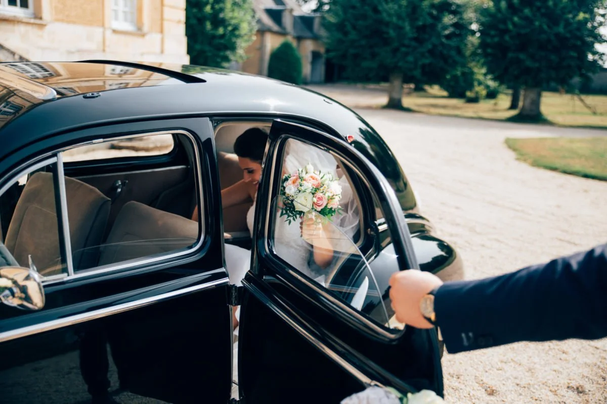 Une mariée tenant un bouquet de fleurs à l'intérieur d'une voiture noire, alors qu'une personne en costume ouvre la portière pour elle.