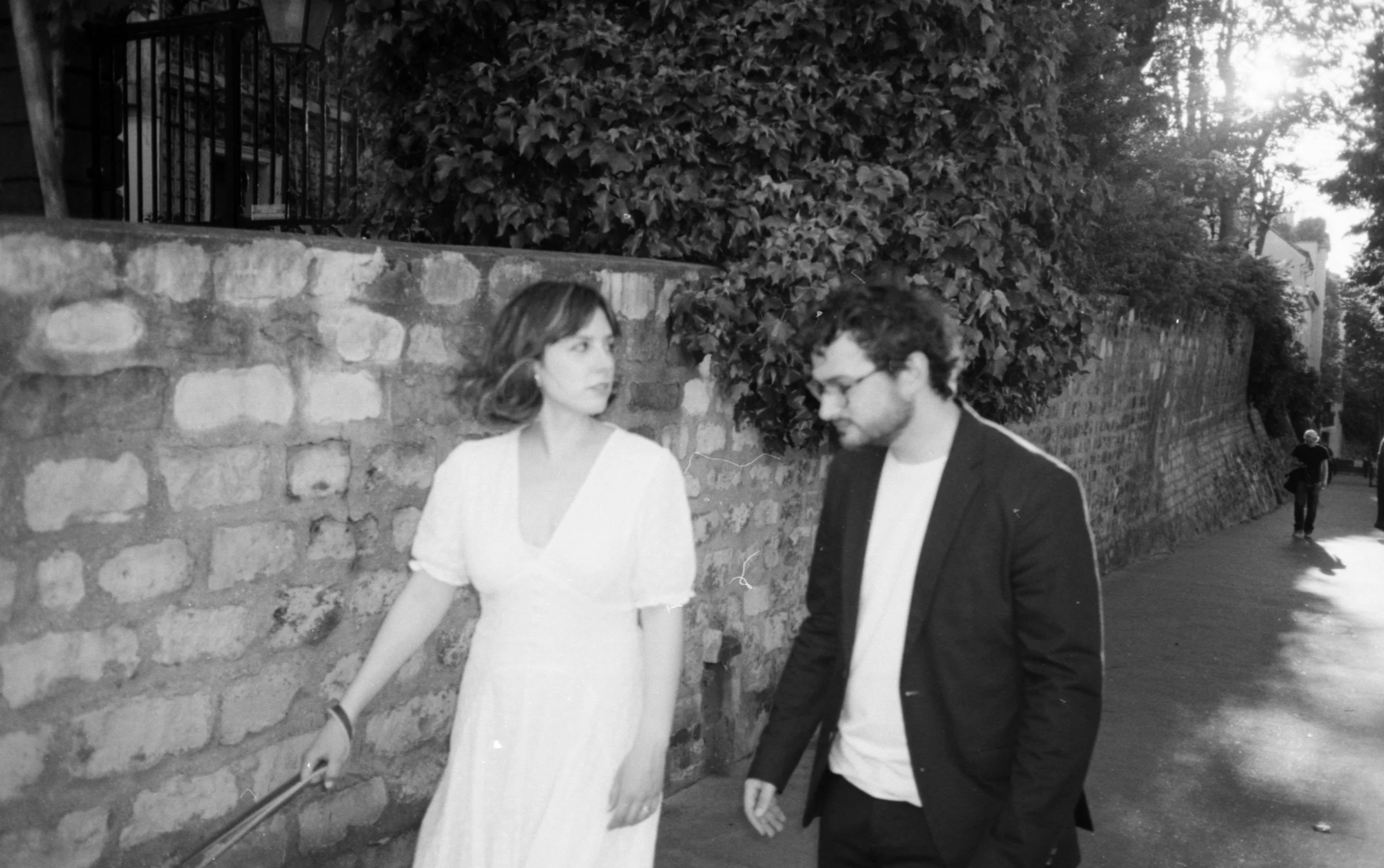 Deux mariés, une femme en robe blanche et un homme en costume sombre, marchent côte à côte dans une rue bordée d'un mur en pierre et de haies verdoyantes, dans un éclairage de fin d'après-midi.