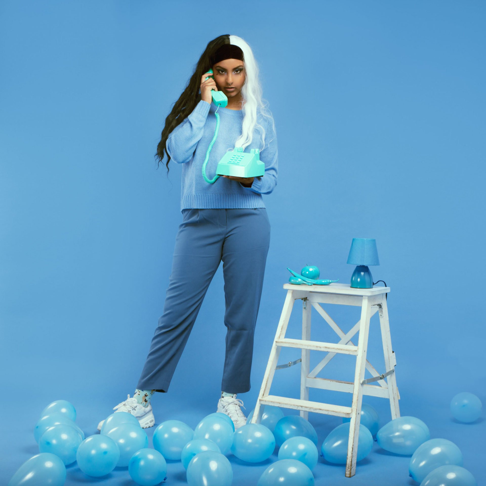 Jeune femme avec cheveux mi-noirs, mi-blancs, portant un pull bleu, parlant au téléphone turquoise, debout parmi des ballons bleus et à côté d'une petite table en bois avec une lampe bleue, contre un fond bleu clair.