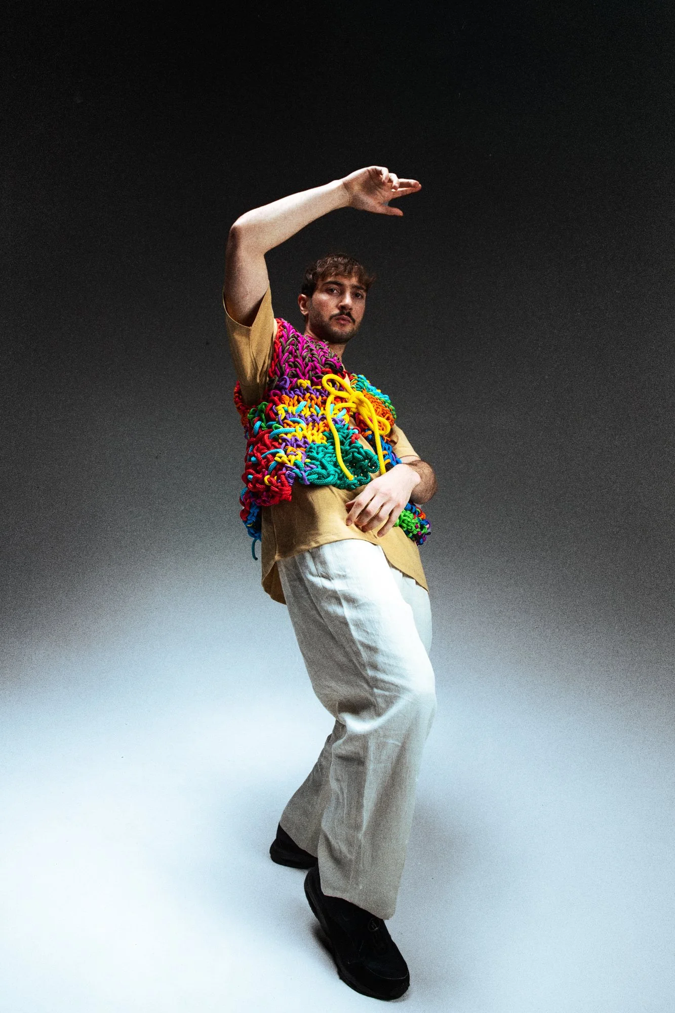 Un homme debout dans un studio sombre, portant un vêtement rayé avec un accessoire en forme de nœud coloré sur le torse, pantalon beige et chaussures noires, positionné de manière dynamique.