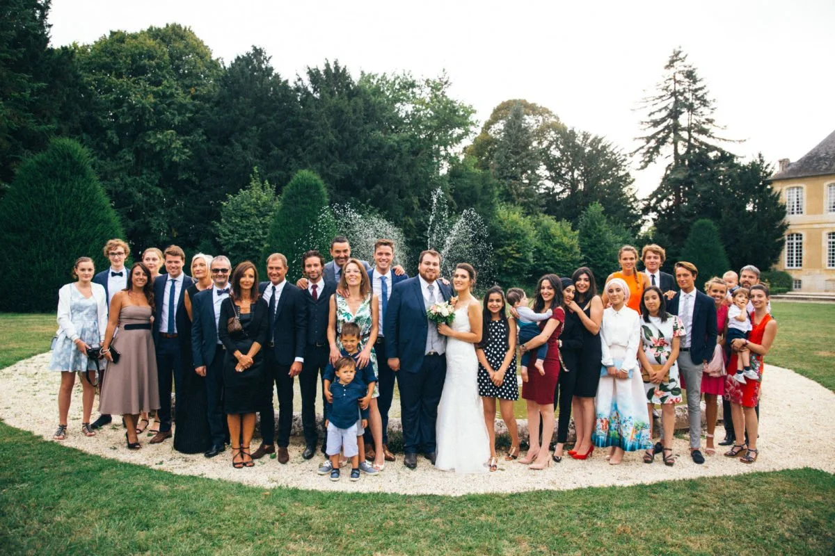 Groupe de personnes en habits de cérémonie lors d'un mariage en extérieur, avec un espace vert et un bâtiment en arrière-plan.