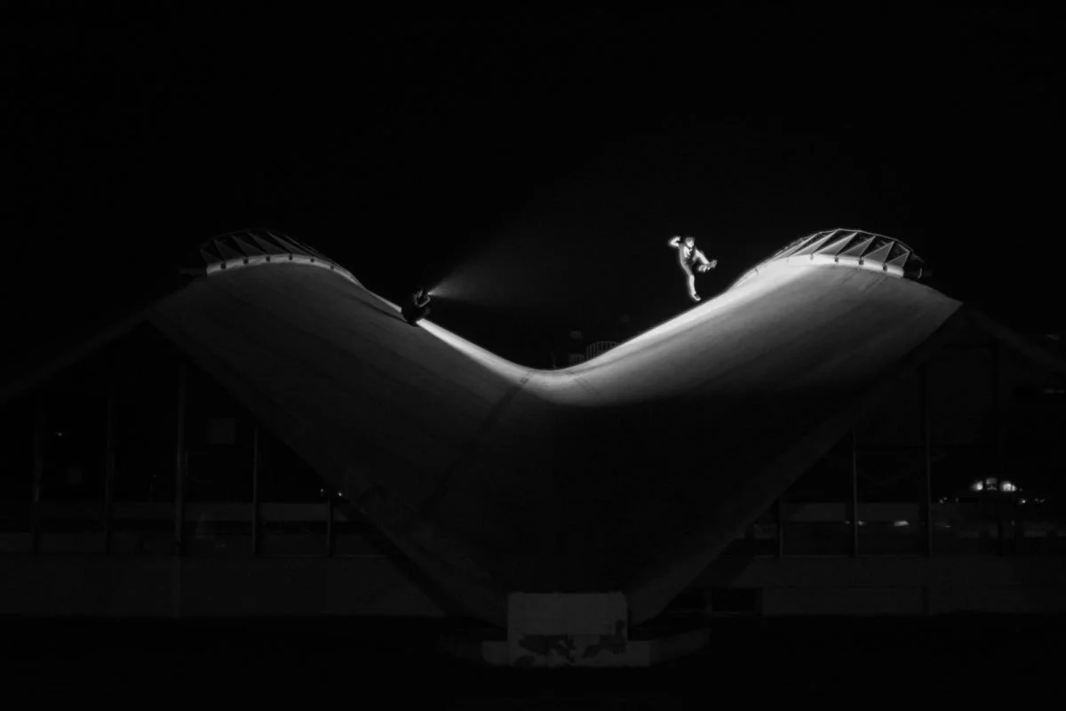 Un skateur effectuant une figure sur un skyline éclairé la nuit, avec un bâtiment en forme de cœur en arrière-plan.