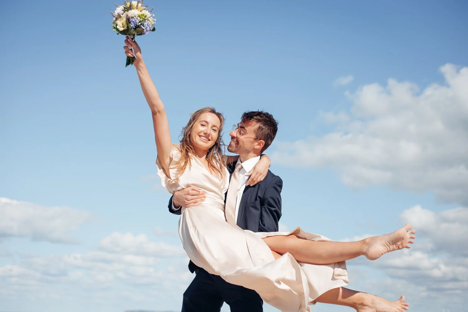 Un homme en costume noir portant une femme en robe blanche, qui tient un bouquet de fleurs, contre un ciel bleu avec quelques nuages, lors d'une célébration de mariage.