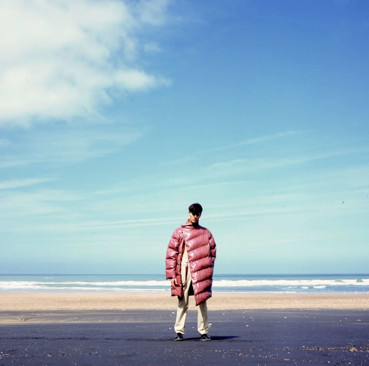 Une personne portant un manteau rembourré rose vif se tient sur une plage au bord de la mer, sous un ciel bleu avec quelques nuages. Arthur Avellano