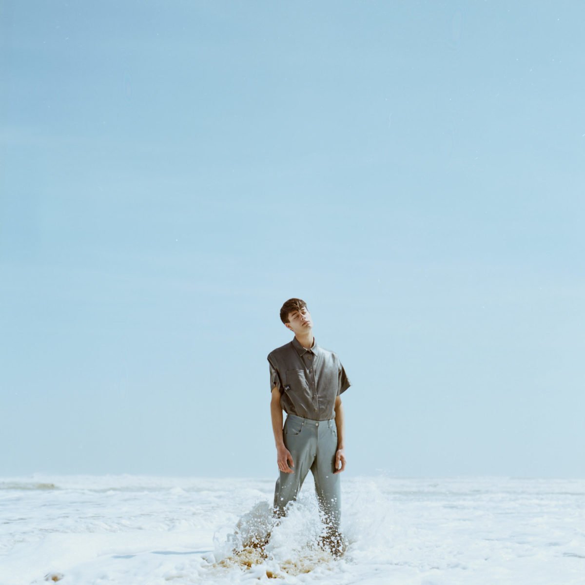 Jeune homme debout dans la mer, portant une chemise grise, sous un ciel bleu clair. Arthur Avellano