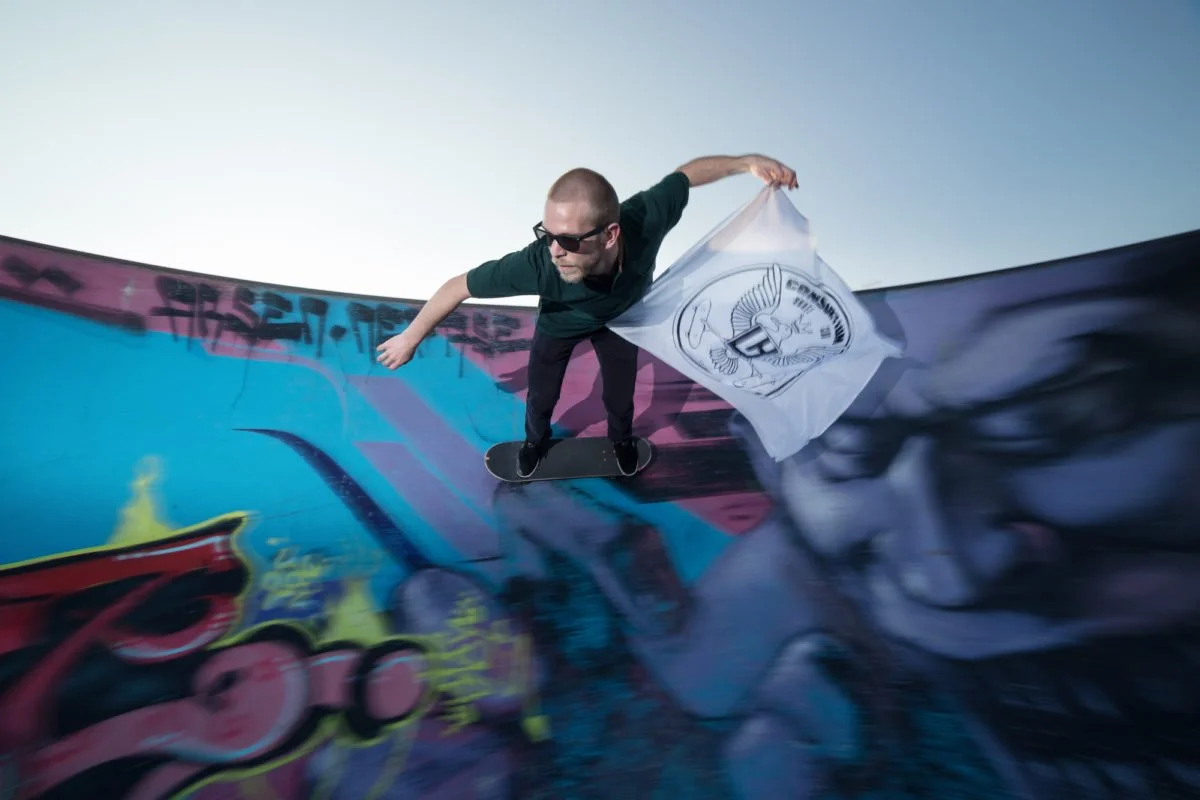 Un homme portant des lunettes de soleil fait du skateboard dans un skatepark au havre avec des graffitis, tout en tenant un drapeau blanc avec un logo d'ailes et un bouclier.