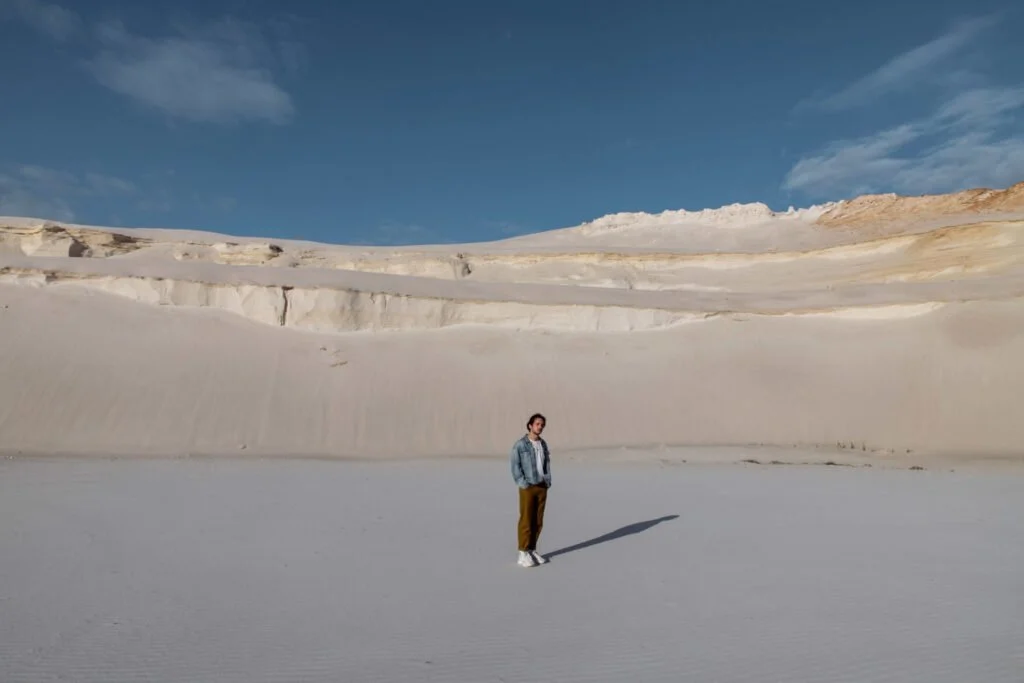 Une personne se tient seule dans un paysage désertique blanc avec des formations rocheuses ou congelées et un ciel bleu clair.