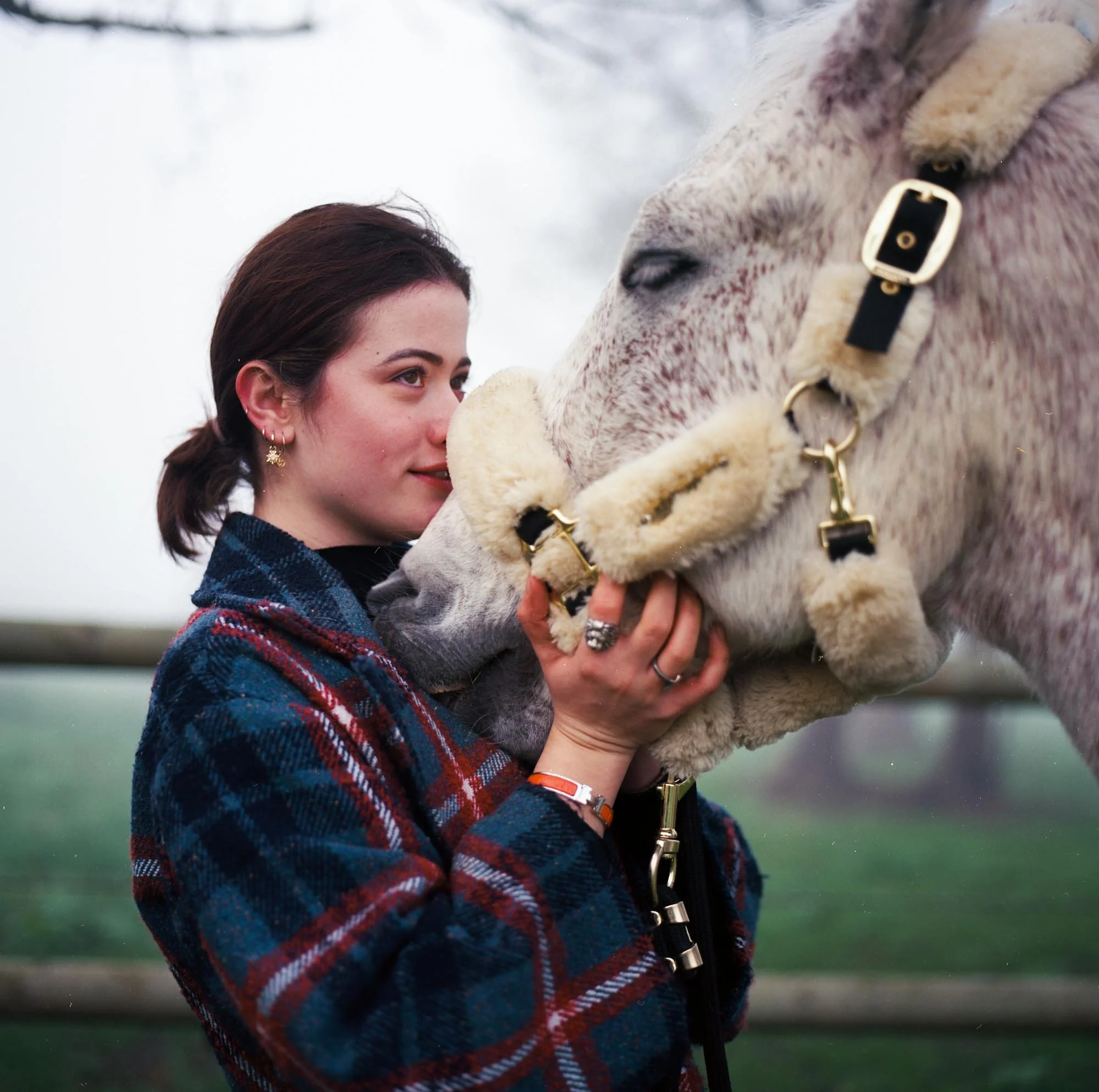 Mathilde & Sligo regarde tendrement un cheval blanc lors d'une journée froide, avec un fond flou de terrain vert et de ciel nuageux.