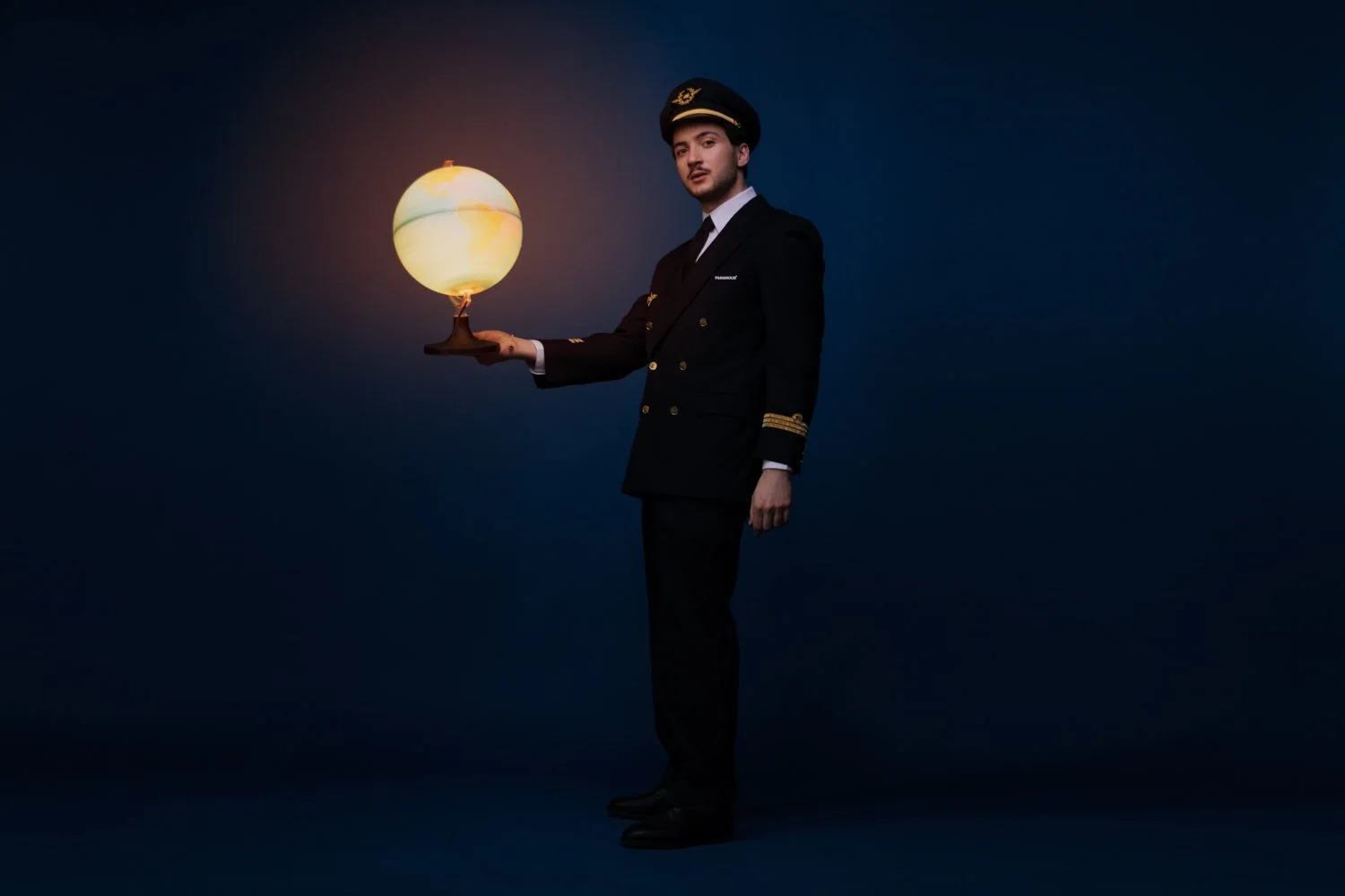Un homme en uniforme de pilote de ligne tient une lampe qui ressemble à une planète ou à un soleil dans un décor sombre.