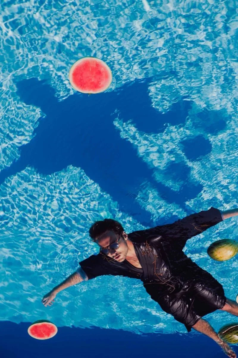 Jeune homme nageant dans une piscine, portant des lunettes de soleil, avec des pastèques en flottants et des ballons d'eau à la surface.