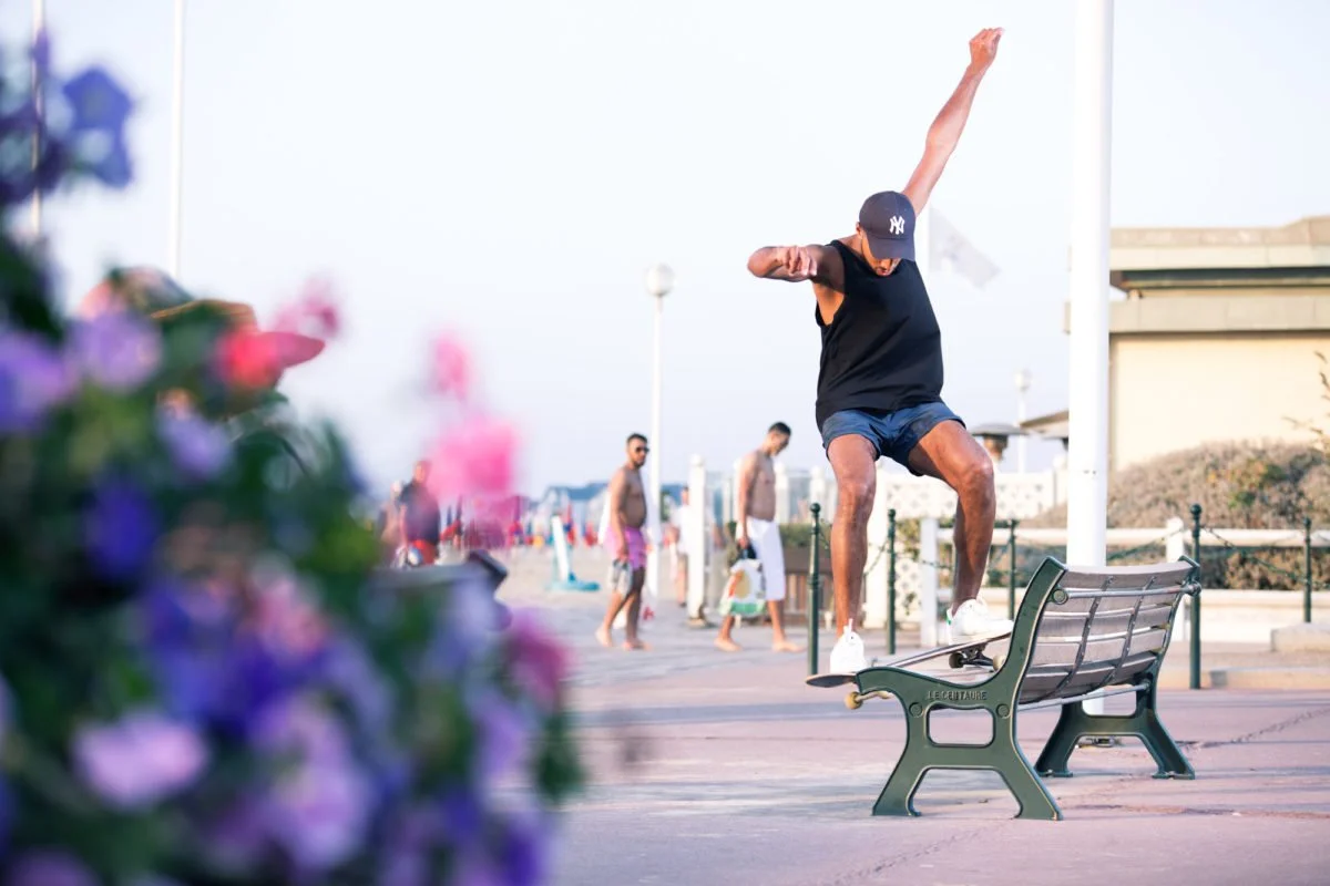 Un jeune homme en débardeur noir, shorts bleus et casquette font du skateboard sur un banc au bord de la plage de Deauville, d'autres personnes se promènent en arrière-plan.