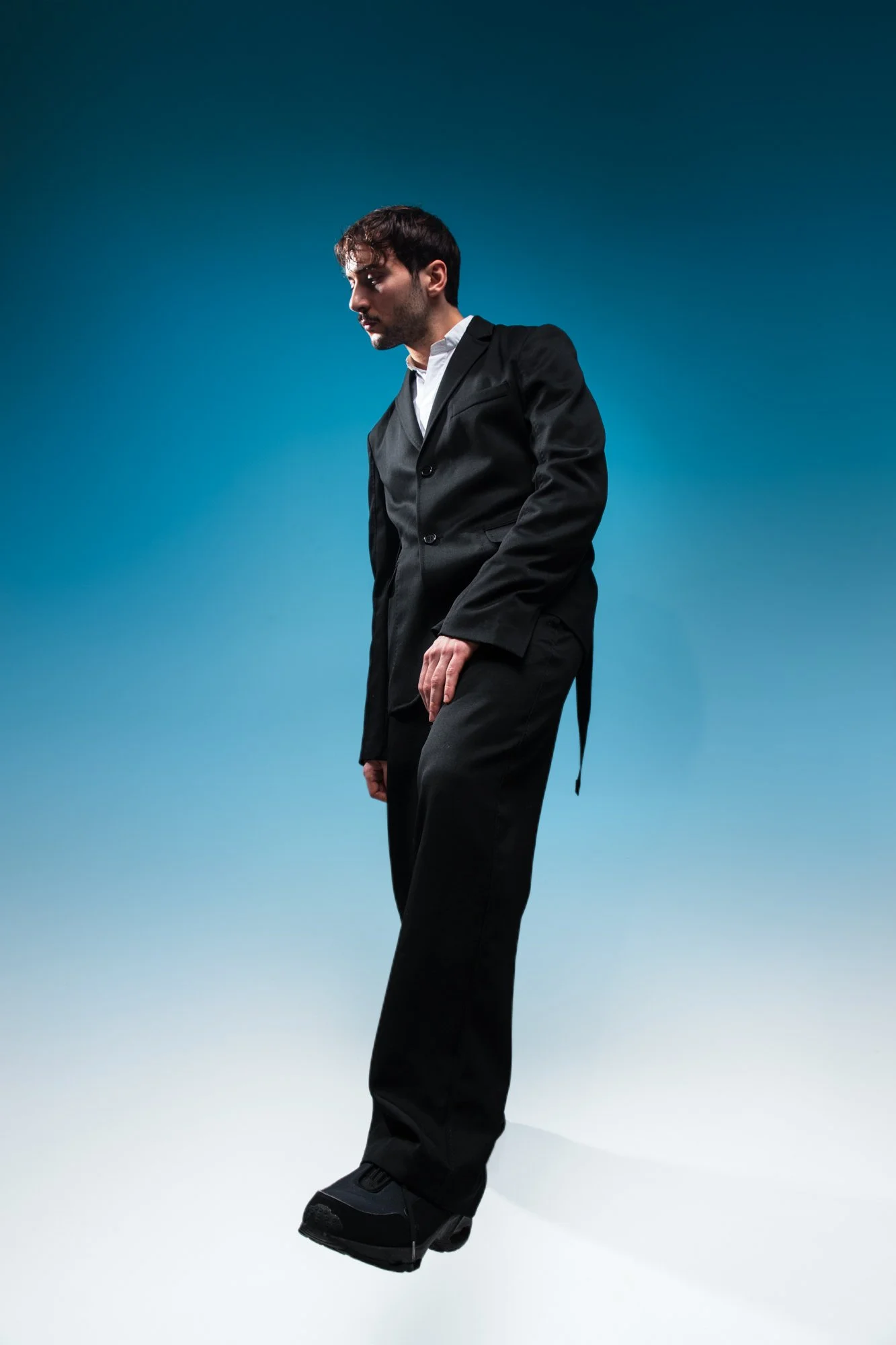 Un homme habillé en costume noir, portant des bottes spéciales, debout dans un studio à fond bleu, donnant l'impression qu'il fait du skateboard ou patine sur le sol.