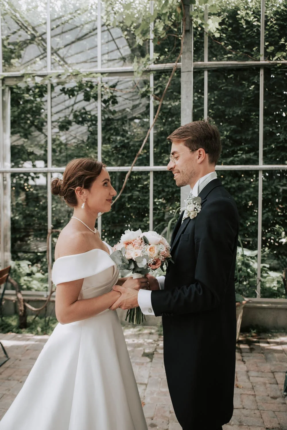 Un couple de mariage échangeant un regard, la mariée portant une robe blanche élégante avec un chignon et des bijoux, le marié en costume noir avec une boutonnière, dans un jardin ou serre avec de la verdure en arrière-plan.