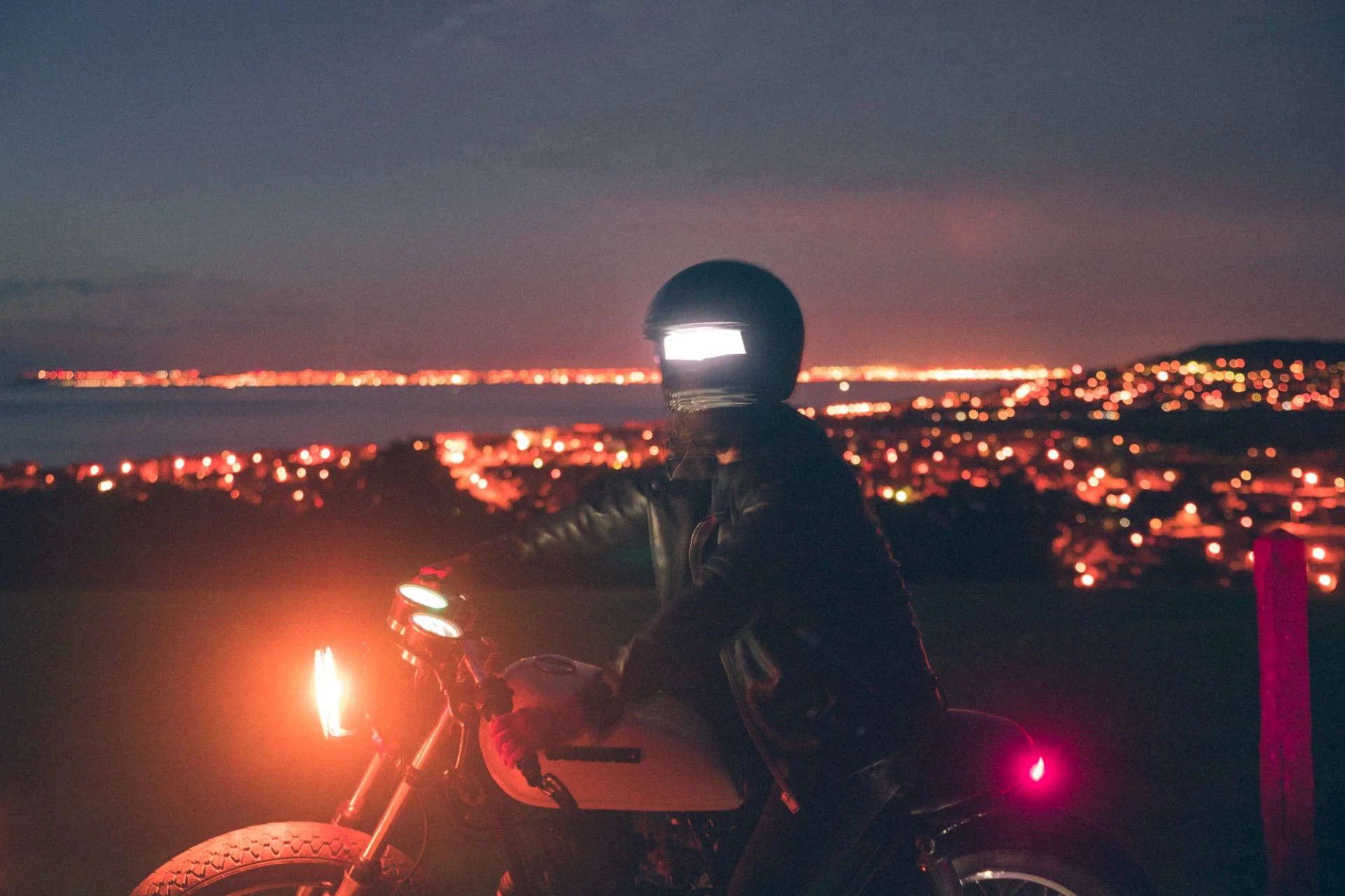 Personne en motocycle avec casque, au crépuscule, vue sur les lumières d'une ville au lointain