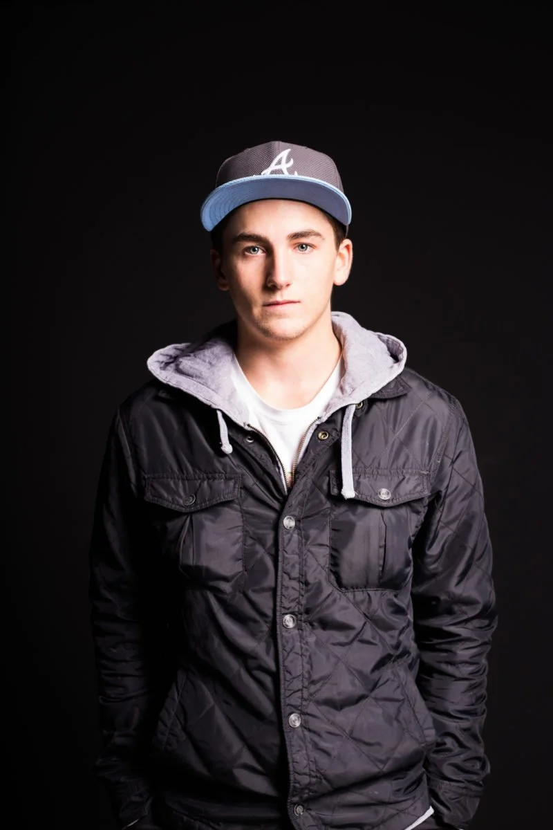 Joseph Garbaccio portant une casquette de baseball grise, une veste noire avec capuche grise et un t-shirt blanc, posé devant un fond noir.