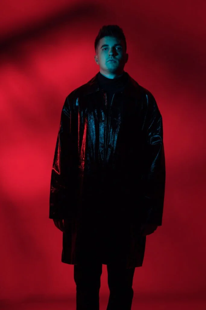 Jeune homme debout portant un manteau en cuir noir sous un éclairage rouge et bleu en arrière-plan.
