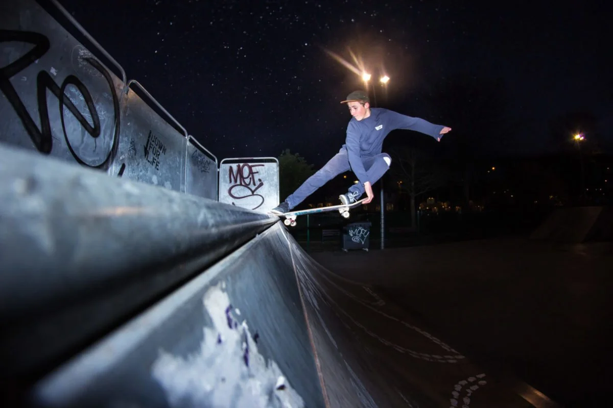 Un skateur en skate effectuant une figure sur un remblai dans un skatepark la nuit, avec des graffitis sur les murs et un ciel étoilé en arrière-plan.