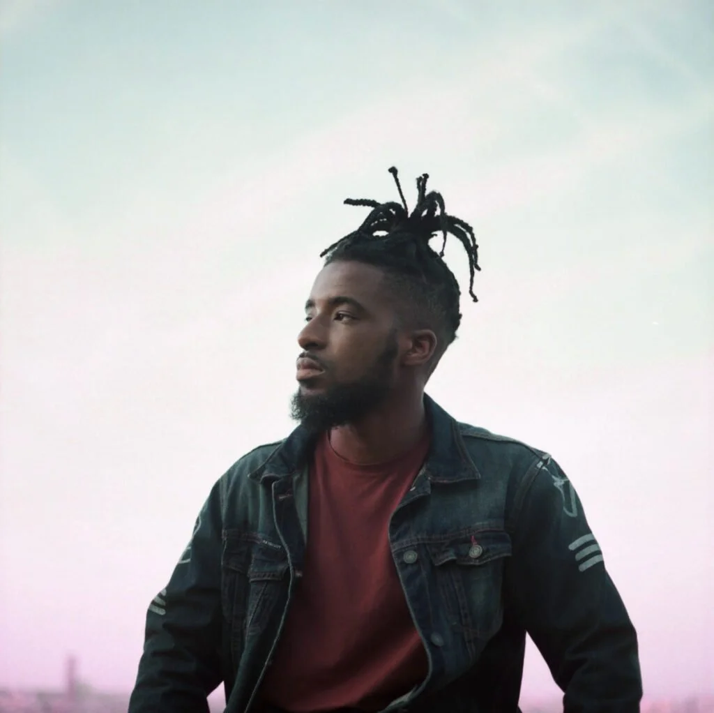 Jeune homme avec dreadlocks, regardant vers la gauche, portant une veste en jean et un t-shirt bordeaux, fond clair.