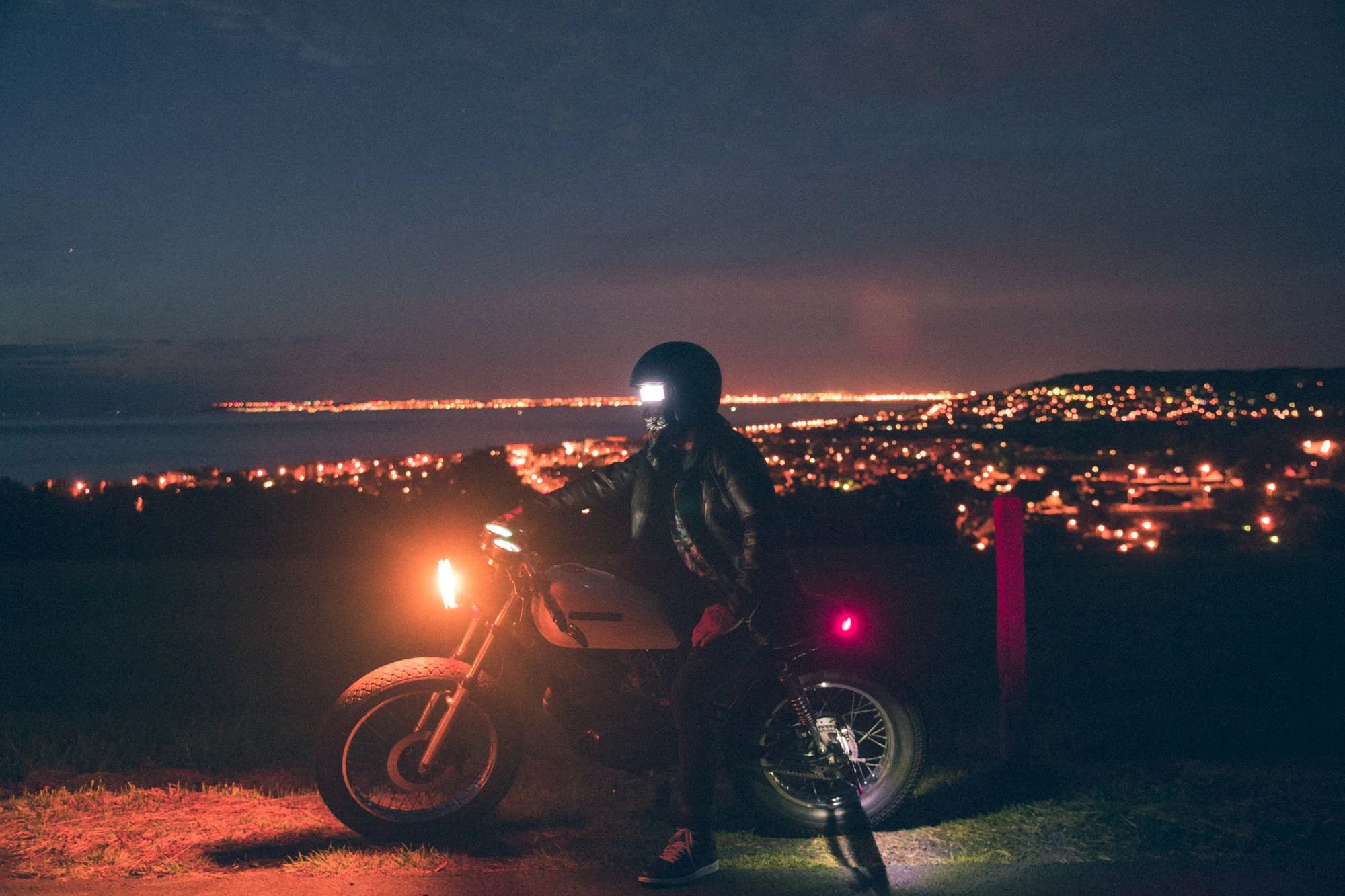 Une personne en casque et vêtement de moto assise sur une moto électrique, au coucher du soleil avec une ville illuminée en arrière-plan et un ciel étoilé.