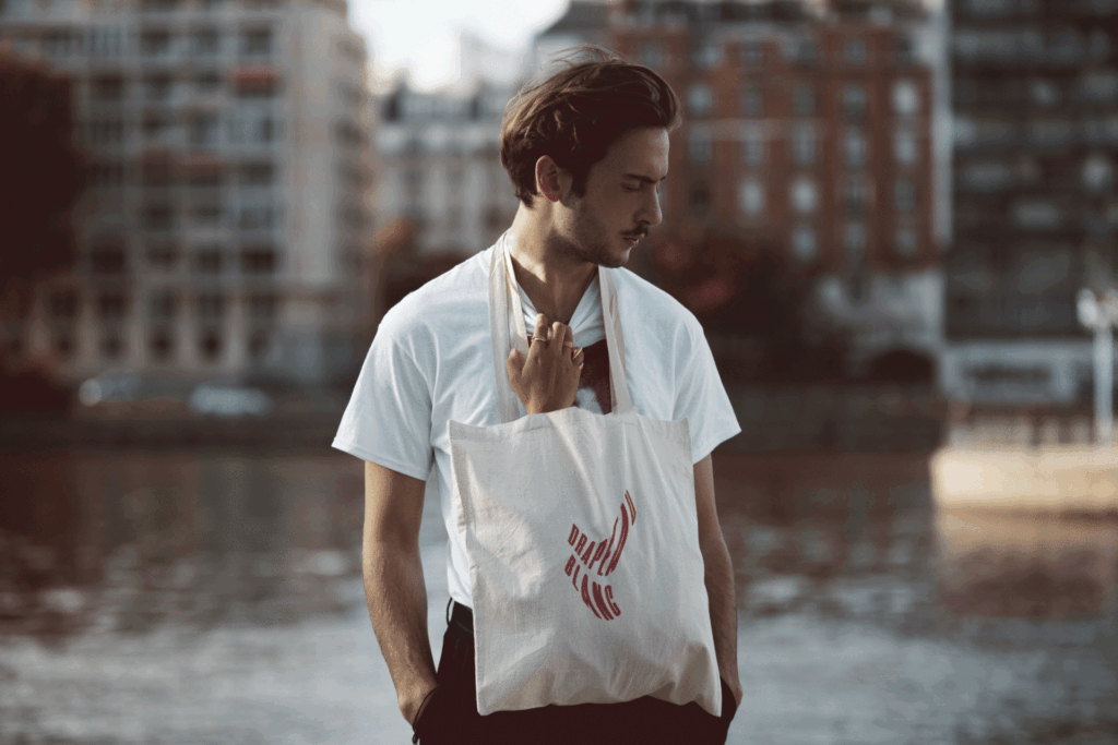 Un jeune homme avec des cheveux bruns, portant une chemise blanche, tenant un sac en toile avec un logo rouge, se tient près d'une rivière en milieu urbain.
