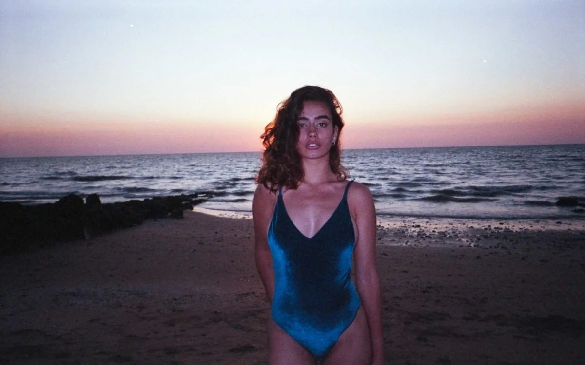 Jeune femme portant un maillot de bain bleu sur la plage au coucher du soleil, avec la mer en fond.