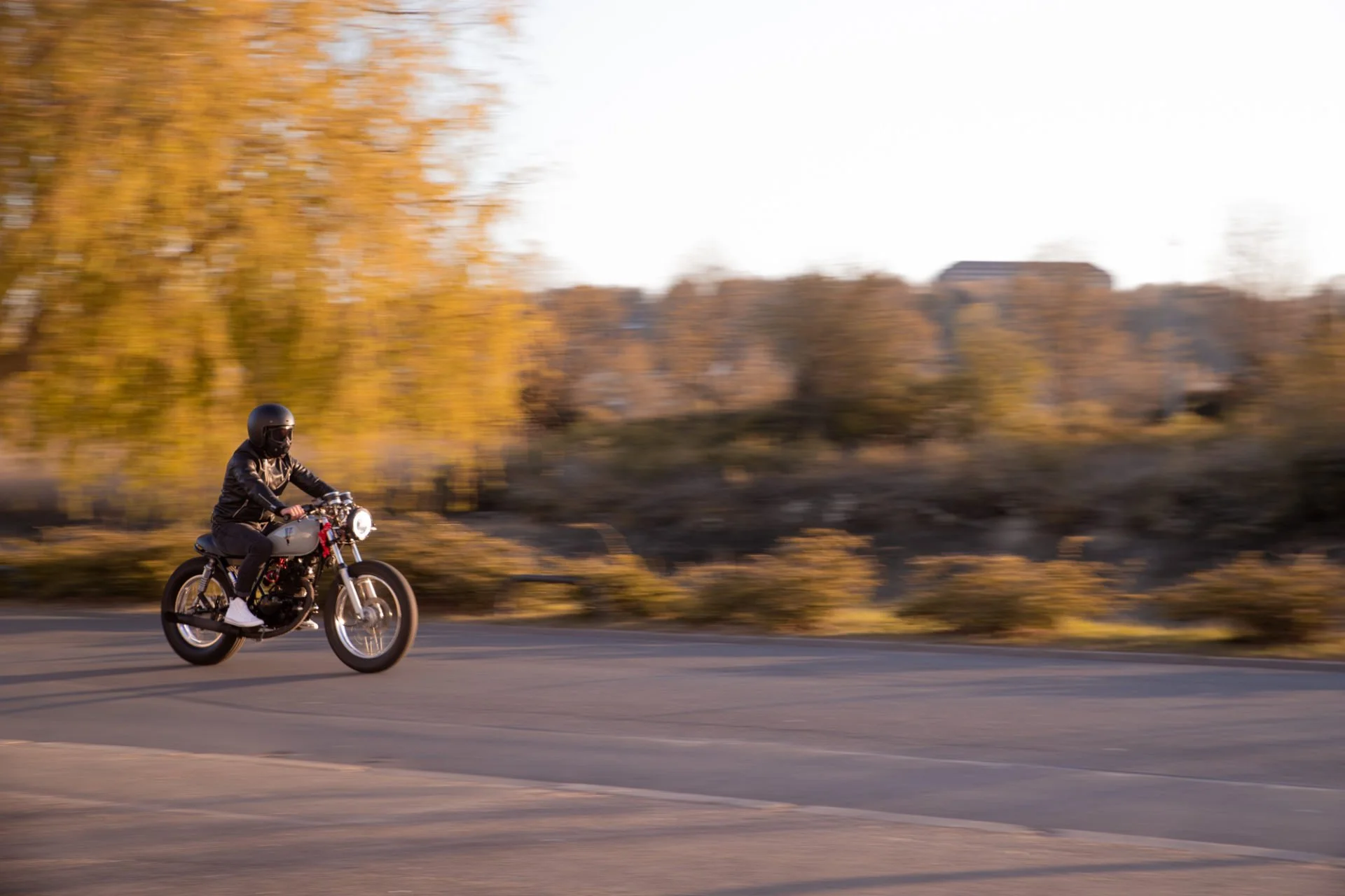 Un homme en casque noir, veste noire, et pantalon noir, conduit une moto blanche à côté d'une route pendant le coucher du soleil, avec des arbres aux feuilles jaunes en arrière-plan.