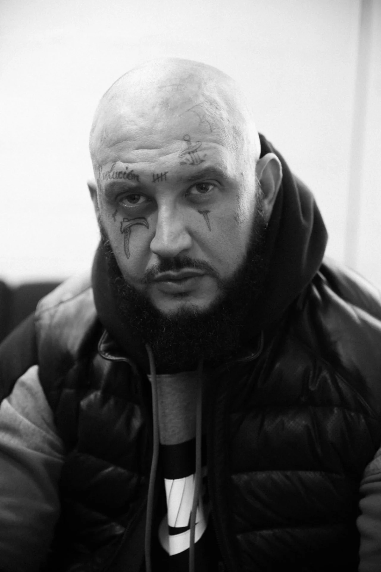 Jeune homme rasé, avec de nombreux tatouages sur le visage, portant une veste en cuir et un hoodie, regardant vers l'objectif.