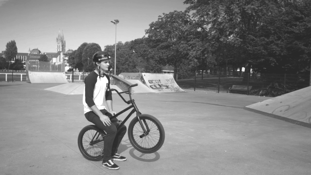 Jeune garçon avec un casque, assis sur son vélo dans un skatepark en plein air, avec des rampes et des arbres en arrière-plan.