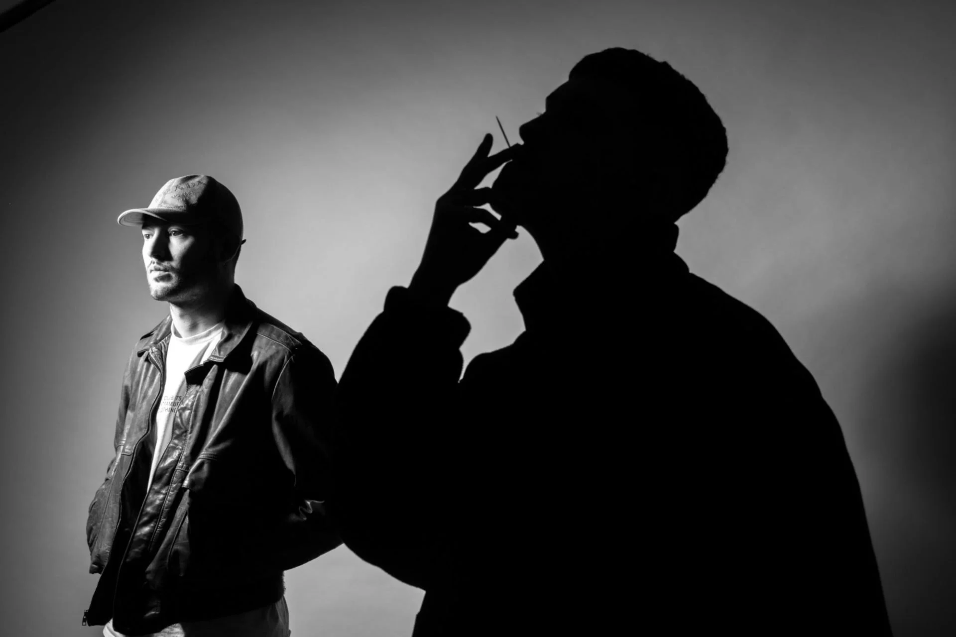 Deux hommes en niveaux de gris, l'un en avant et l'autre en silhouette sombre, avec un fond neutre. L'homme à gauche porte une casquette et une veste en cuir, regardant à gauche, tandis que l'autre homme, en silhouette, a la main près de son visage.
