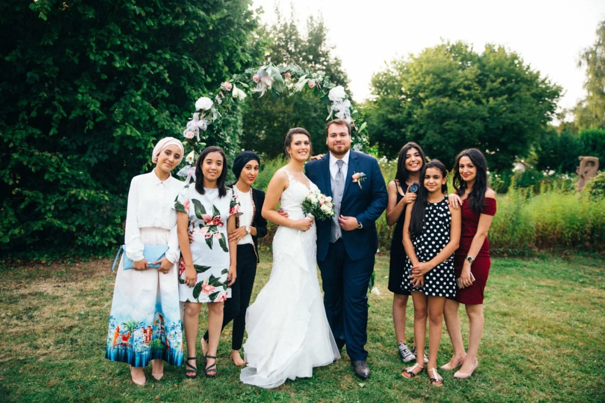 Un groupe de huit personnes — un couple en vêtements de mariage et six autres jeunes femmes — posent dehors sur un terrain herbeux sous un arc fleuri. Le couple est au centre, avec la mariée en robe blanche et le marié en costume bleu.