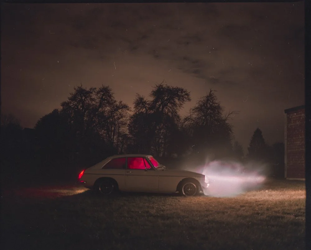 Une voiture ancienne stationnée dans un endroit sombre avec des arbres en arrière-plan, éclairée par les phares allumés et une lumière rougeâtre à l'intérieur.