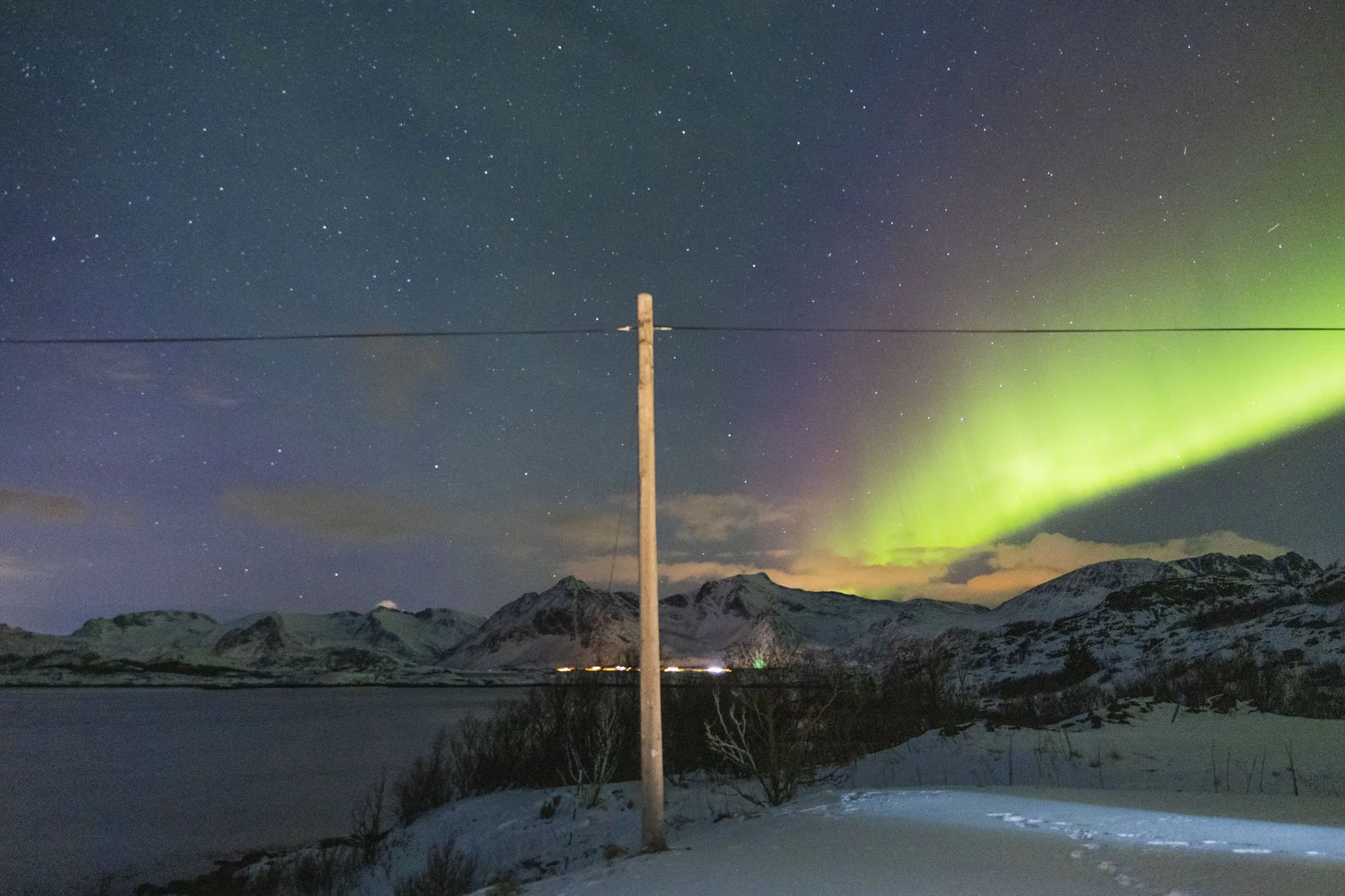 Nuit étoilée avec aurore boréale verte derrière des montagnes enneigées, avec un poteau électrique au premier plan à Tromsø

