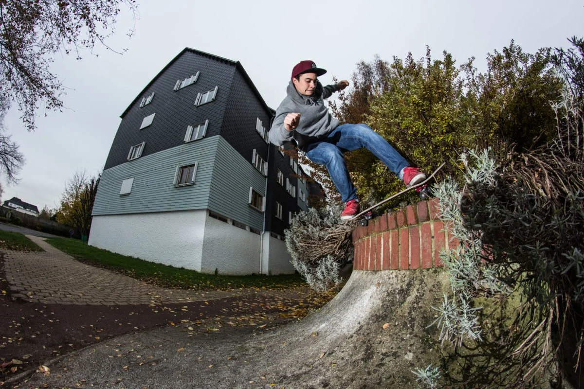 Un jeune homme fait du skateboard sur un petit muret en pierre et briques, avec un bâtiment moderne en arrière-plan, en sous-bois.